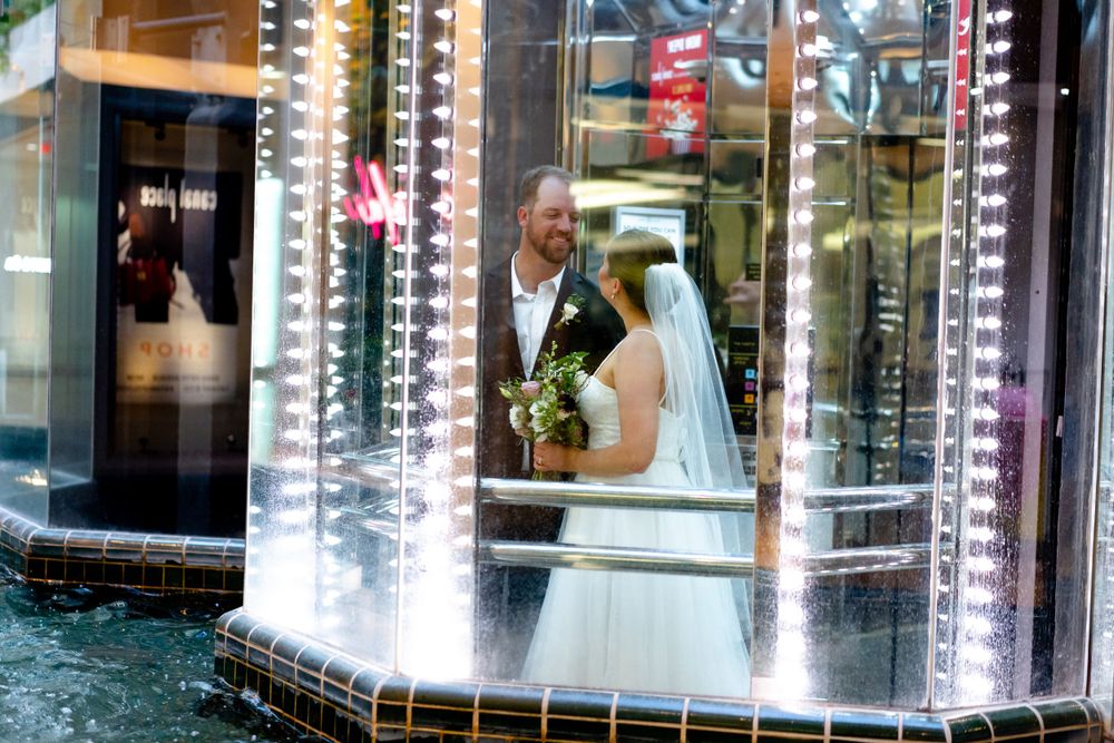 A couple in wedding attire stands in an elevator with glass windows everywhere.