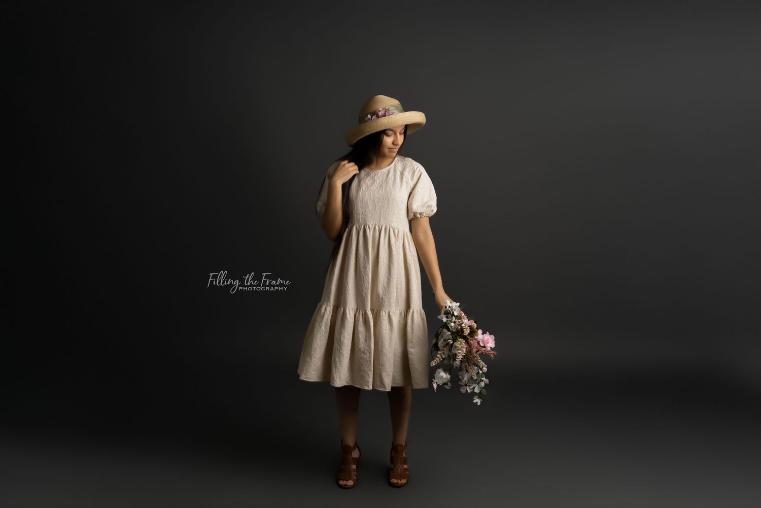 A minimalist portrait against dark background featuring a beige dress and straw hat.