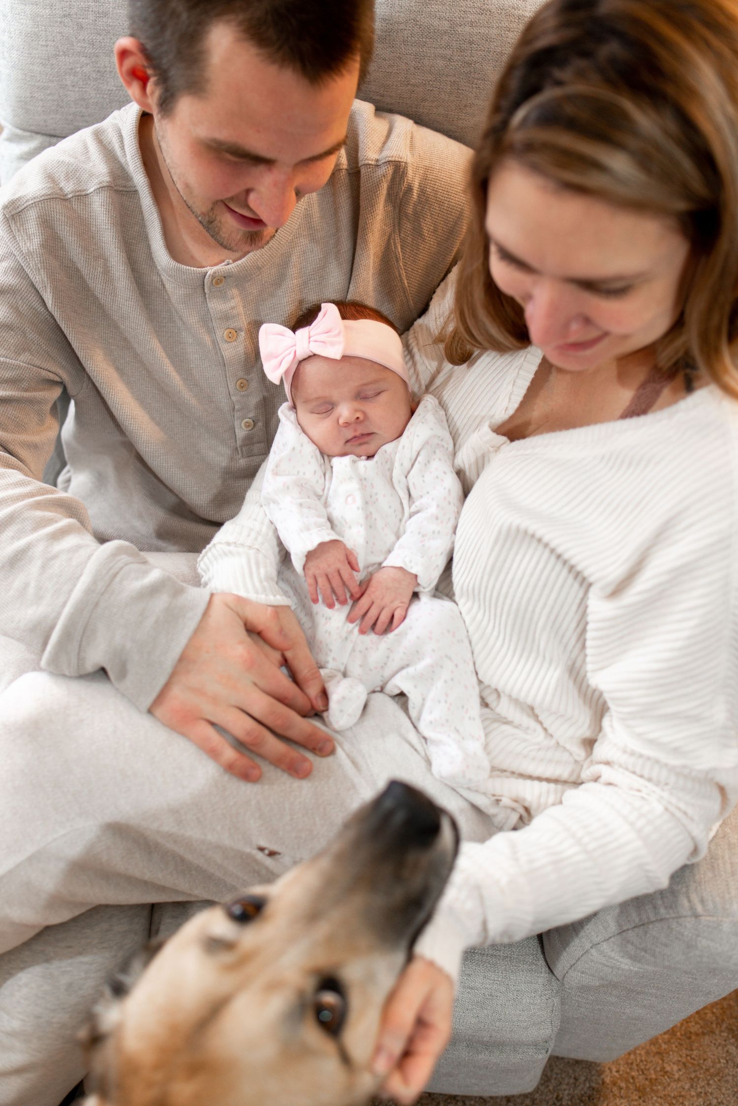 A newborn baby rests peacefully on a bed while being lovingly watched by adults and a dog.