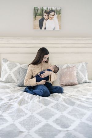 Parent in beige sweater holds baby on bed with framed photo visible on wall.