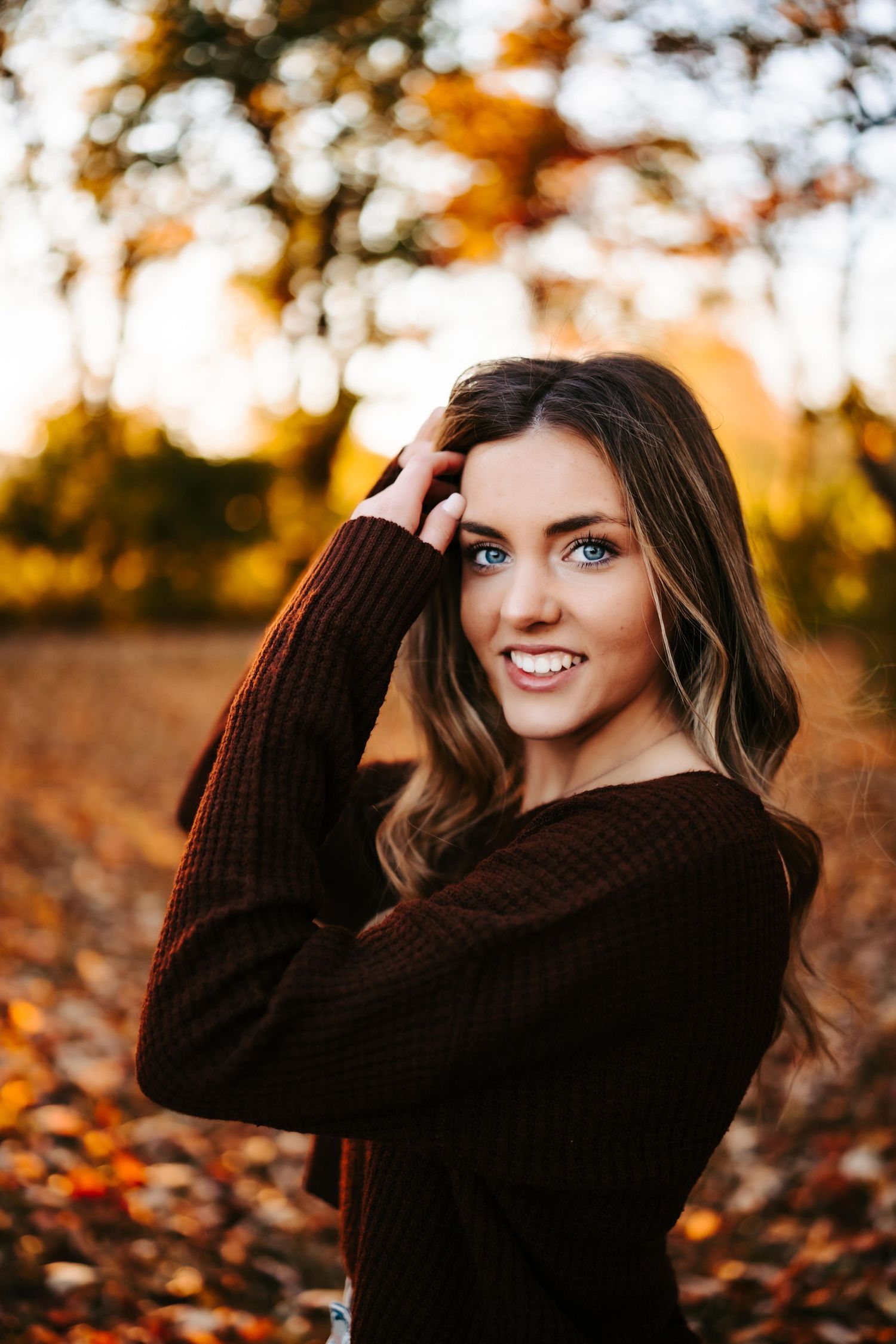Smiling woman with long hair in autumn setting, wearing a dark sweater against a blurred background.