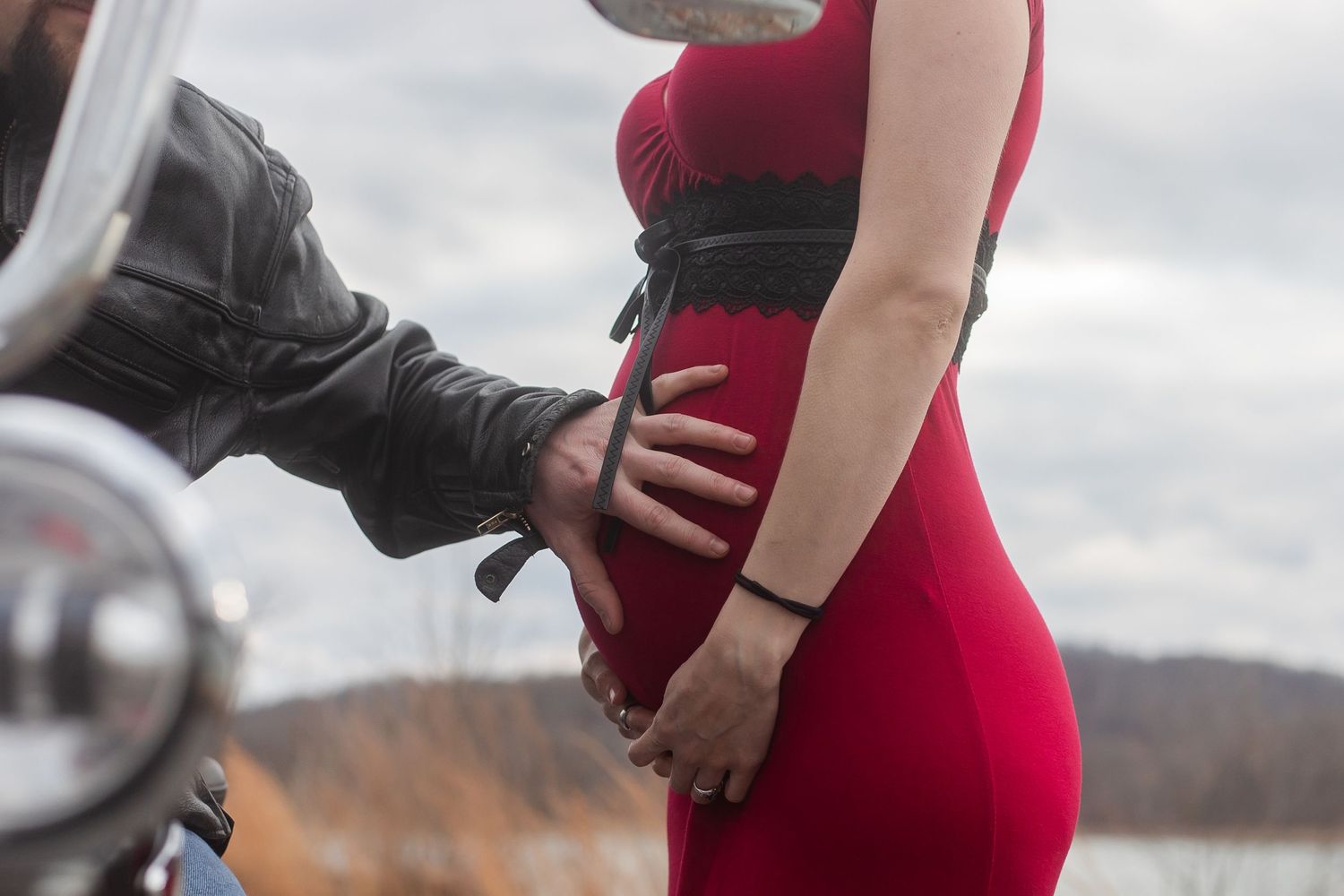 A person in a red dress poses next to a motorcycle by a lake.