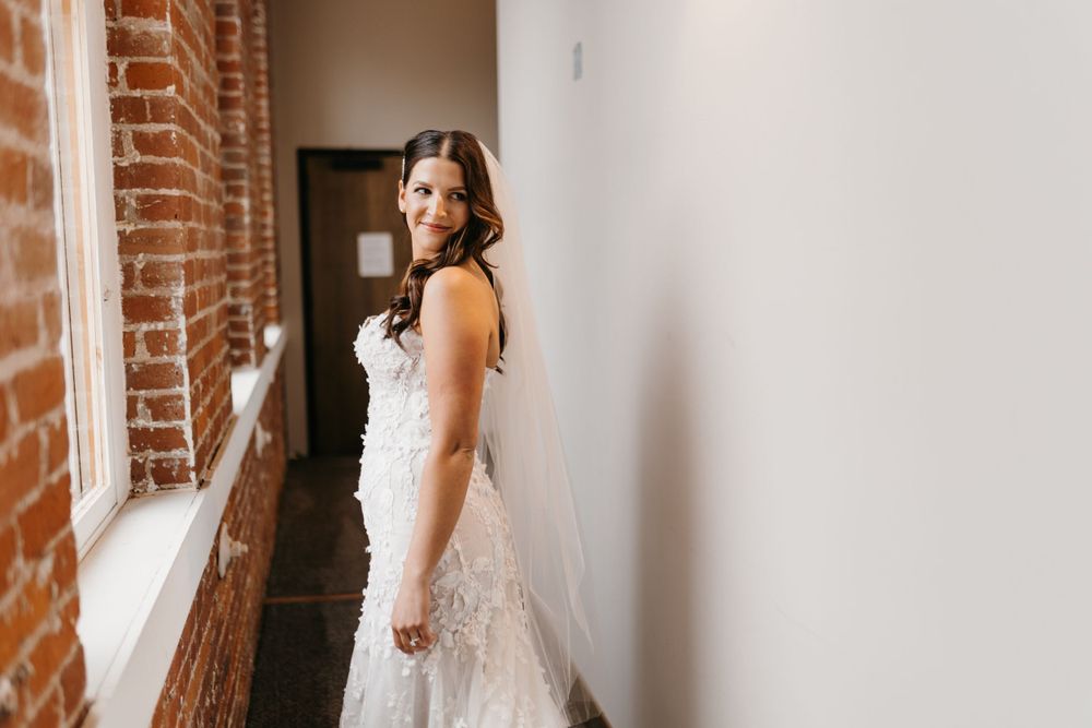 Bride in strapless lace wedding dress smiling over shoulder in hallway with exposed brick wall.