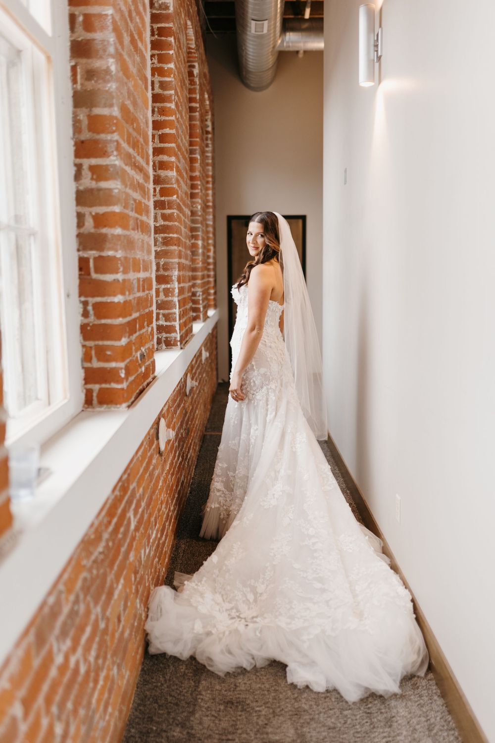 Bride in lace mermaid gown with veil posing in hallway with exposed brick walls and large windows.