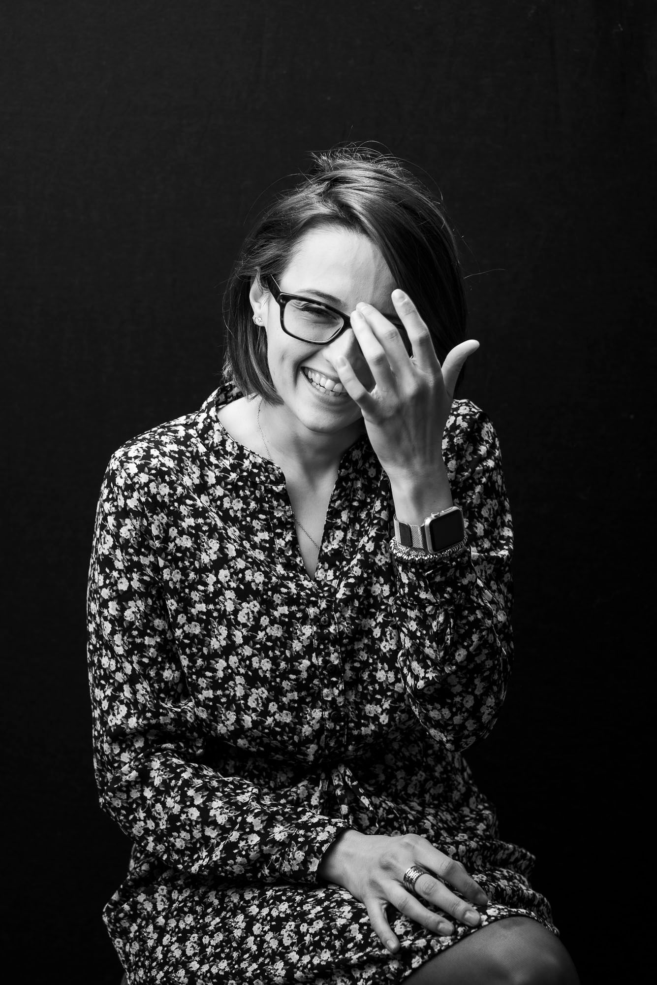 Black and white portrait of a person laughing and covering their face while wearing glasses and a floral print dress.
