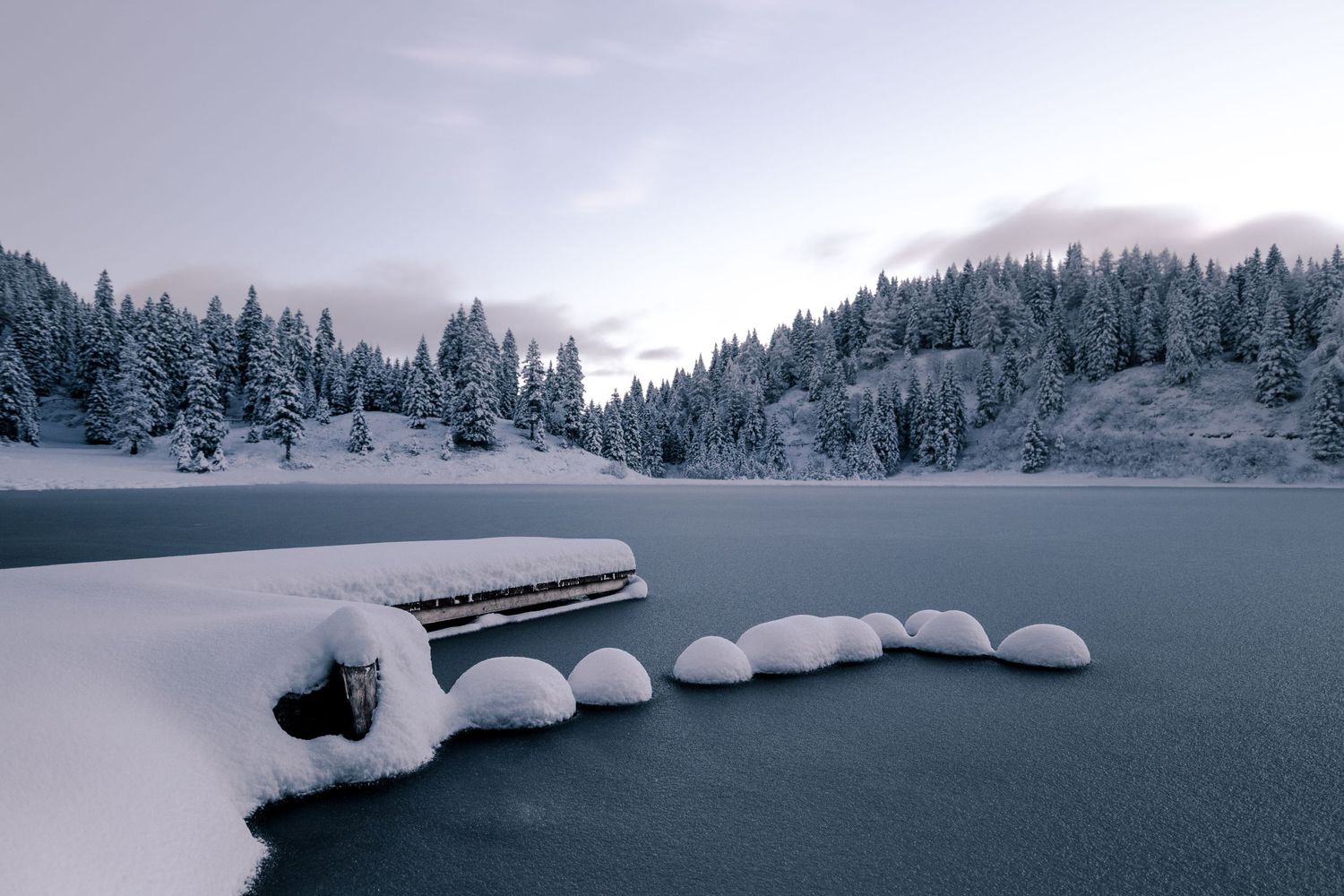 Snow-covered bench by frozen lake surrounded by winter forest in moody lighting.