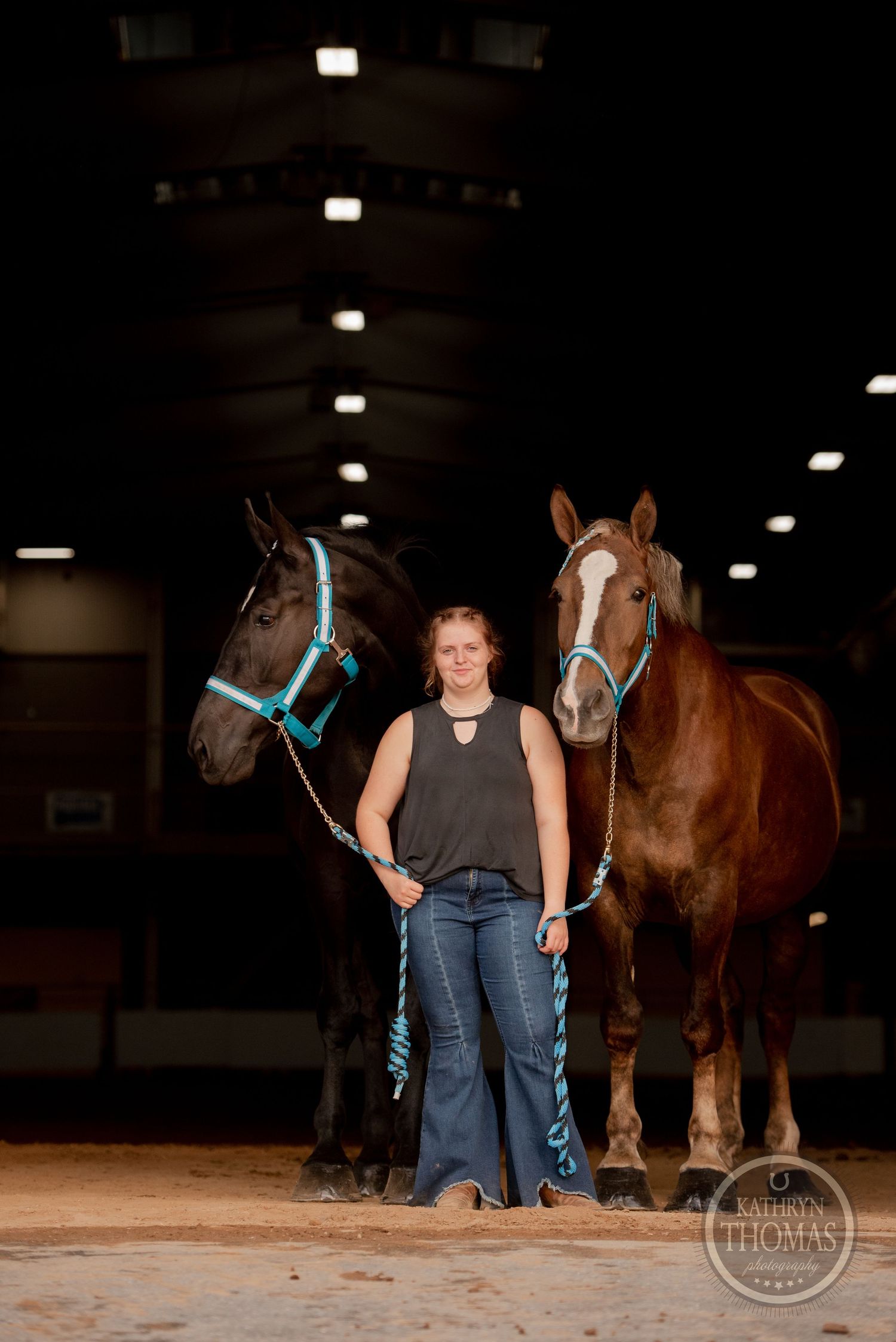 Two horses, one dark and one bay, pose with their handler in an indoor arena.
