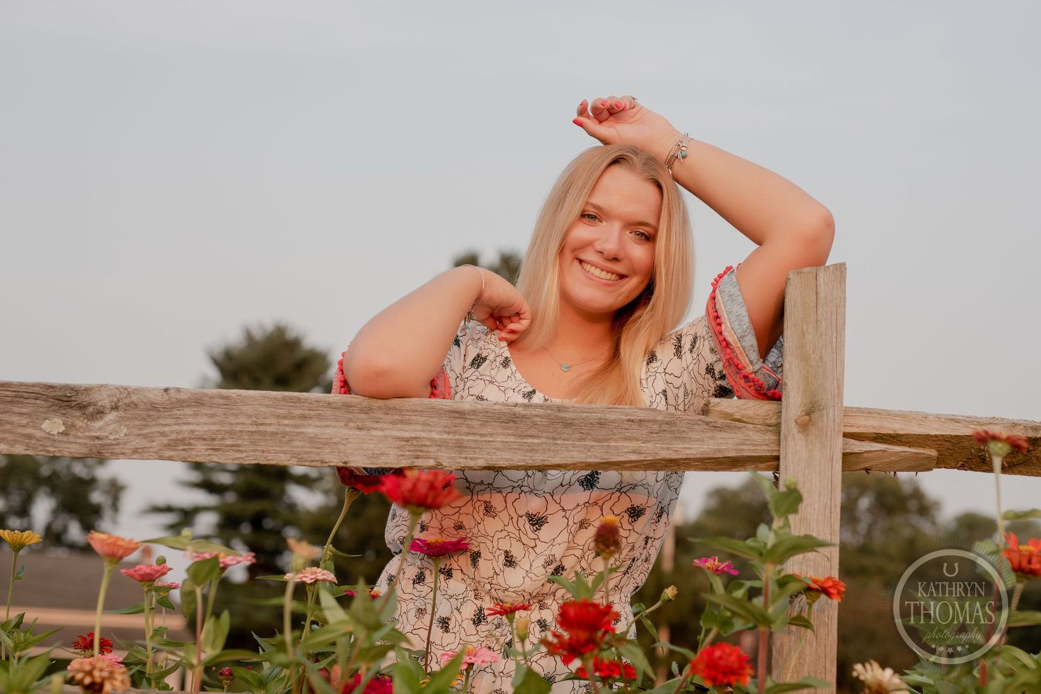 A woman poses playfully at a rustic fence with red flowers in the foreground.