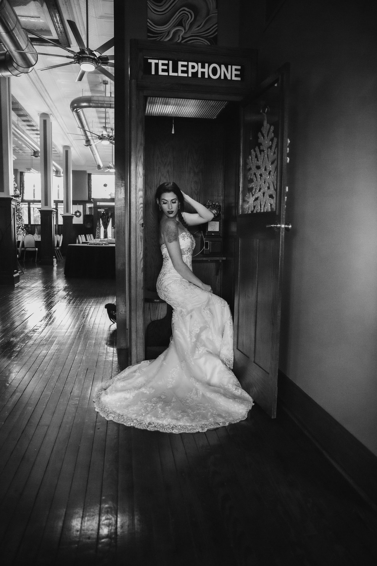 Elegant bride in lace gown poses by vintage telephone booth in classic black and white photo.