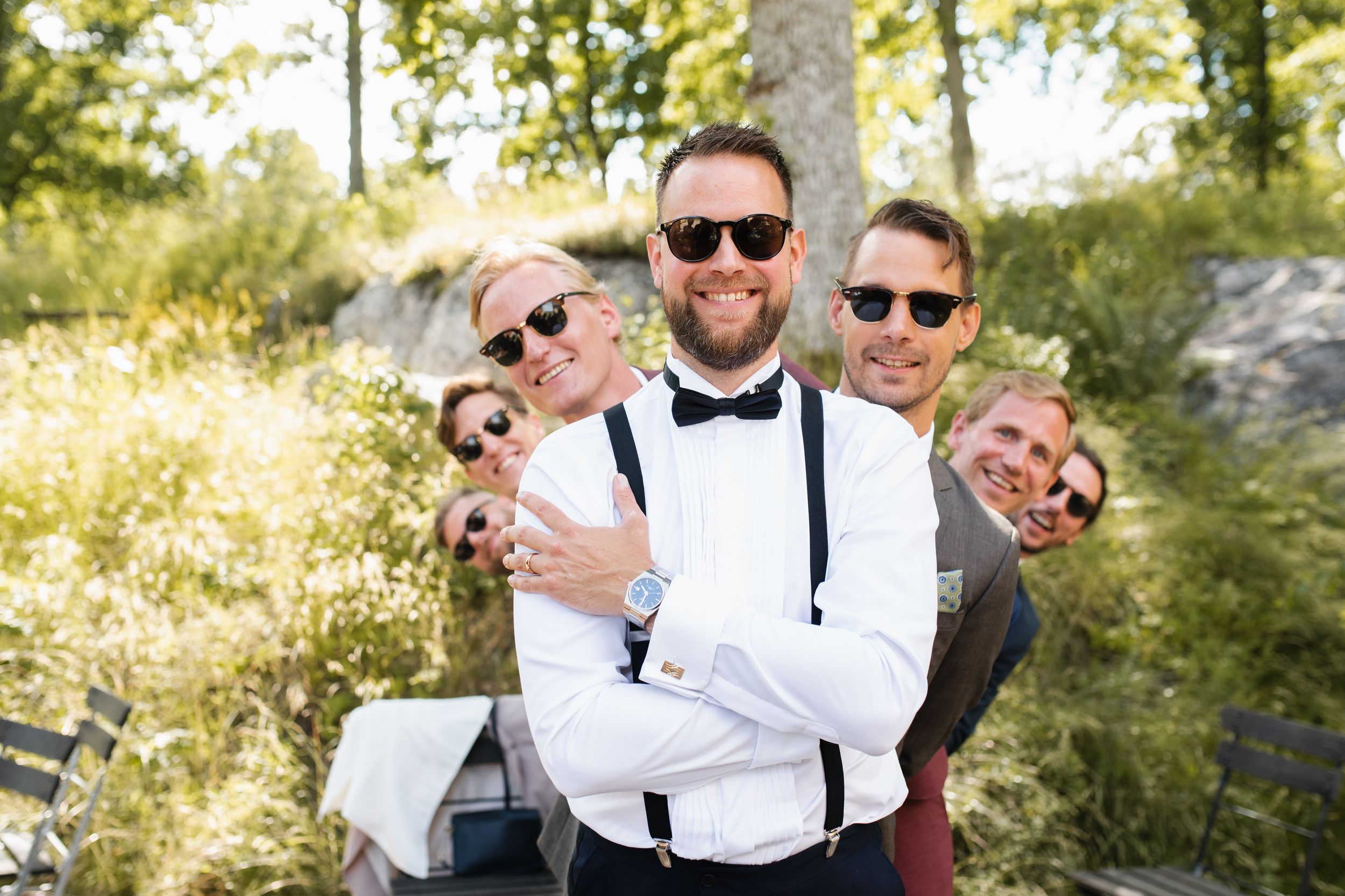 Playful wedding party group poses outdoors with suspenders and sunglasses in natural lighting.