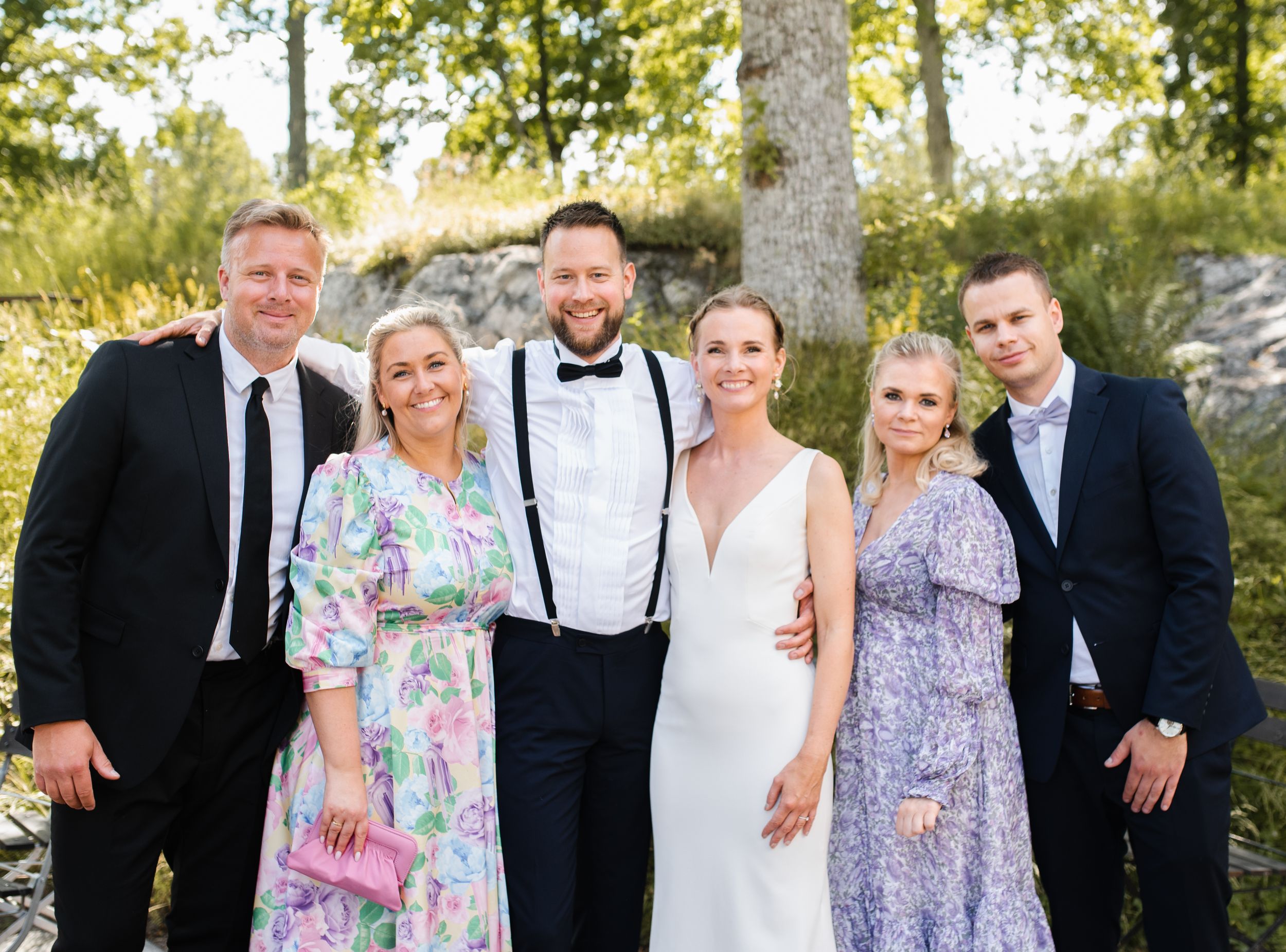 Elegant wedding party poses together outdoors in formal attire against a wooded backdrop.