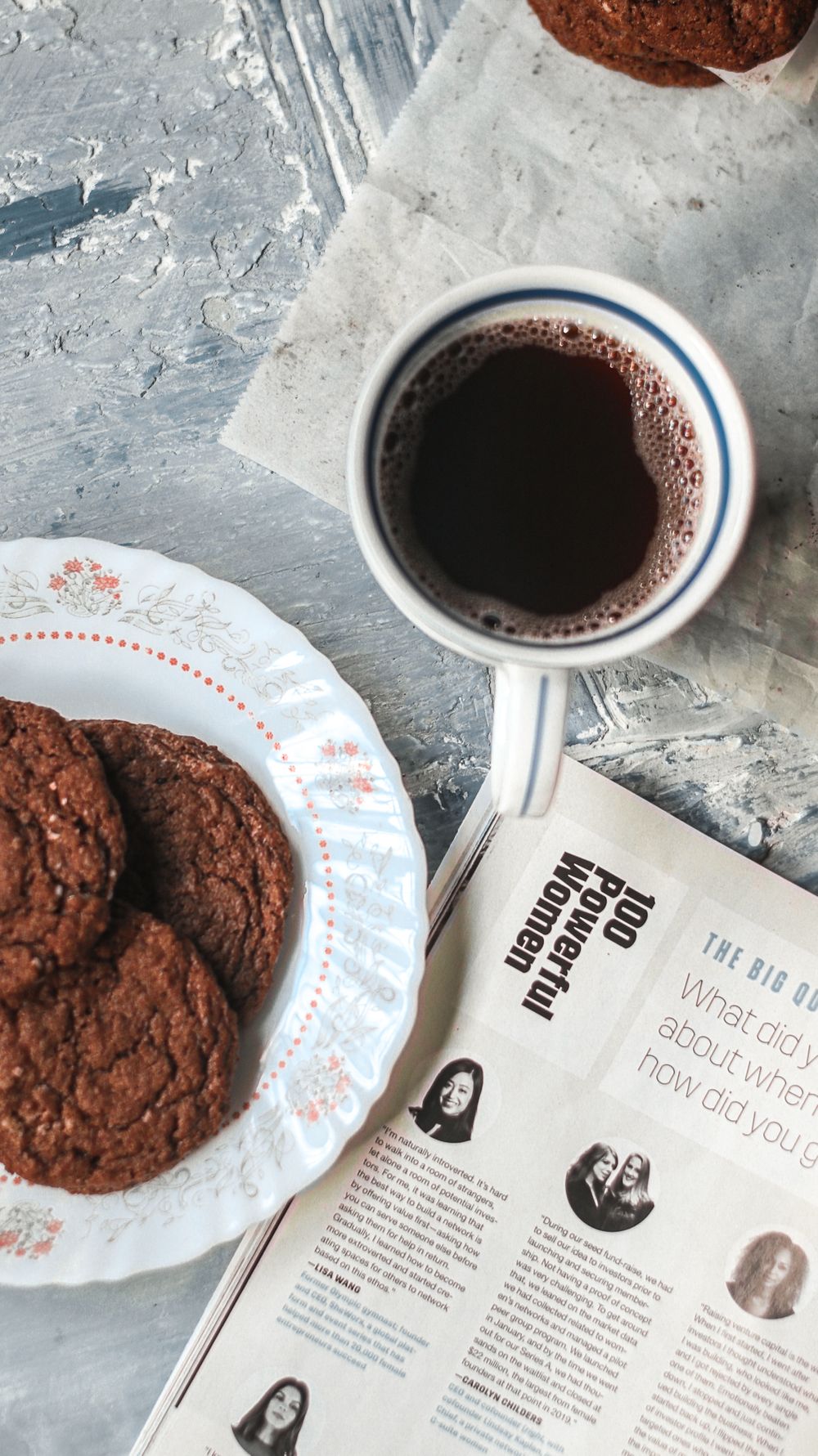 Chocolate cookies on a decorative plate with hot cocoa and a magazine on a textured surface.