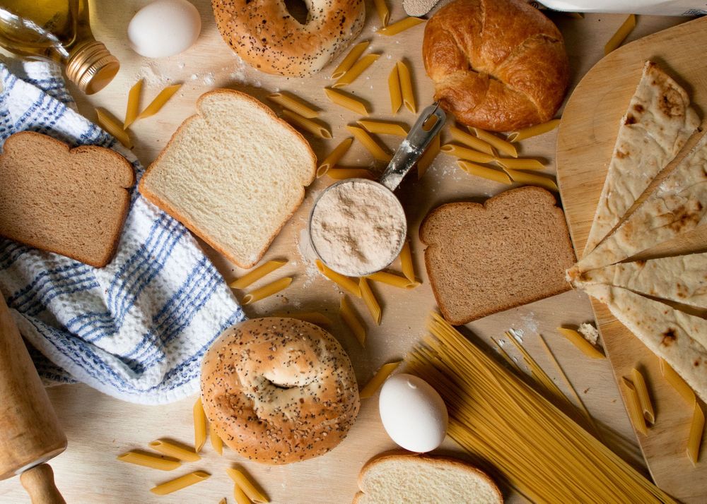 Overhead view of various pasta types, bread, and ingredients arranged on a rustic wooden surface.
