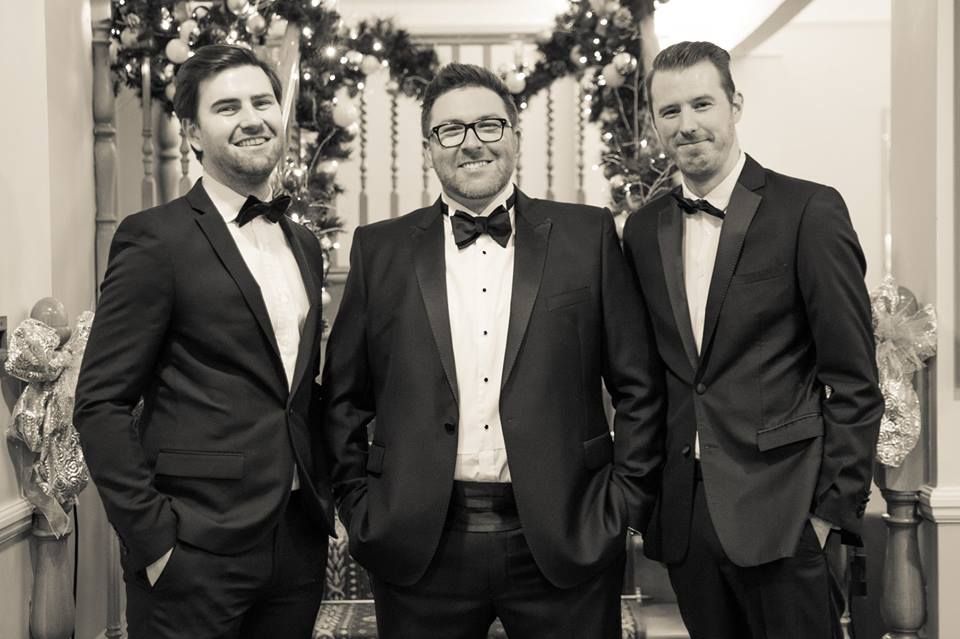 Three men in formal tuxedos pose together in front of holiday decorations.