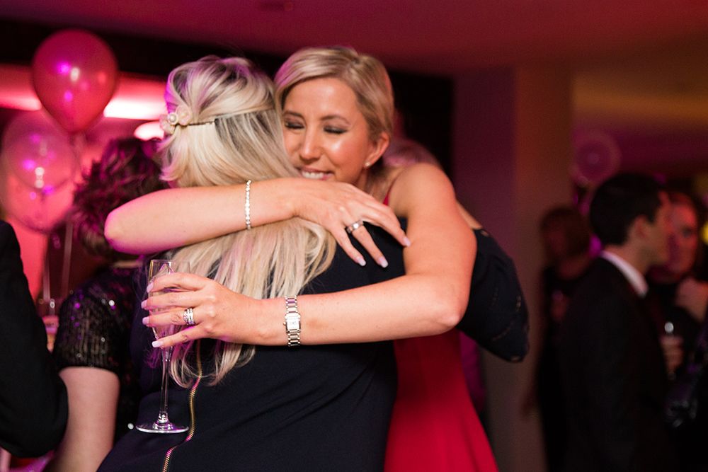 Two partygoers share an emotional embrace during a festive celebration with red balloons.