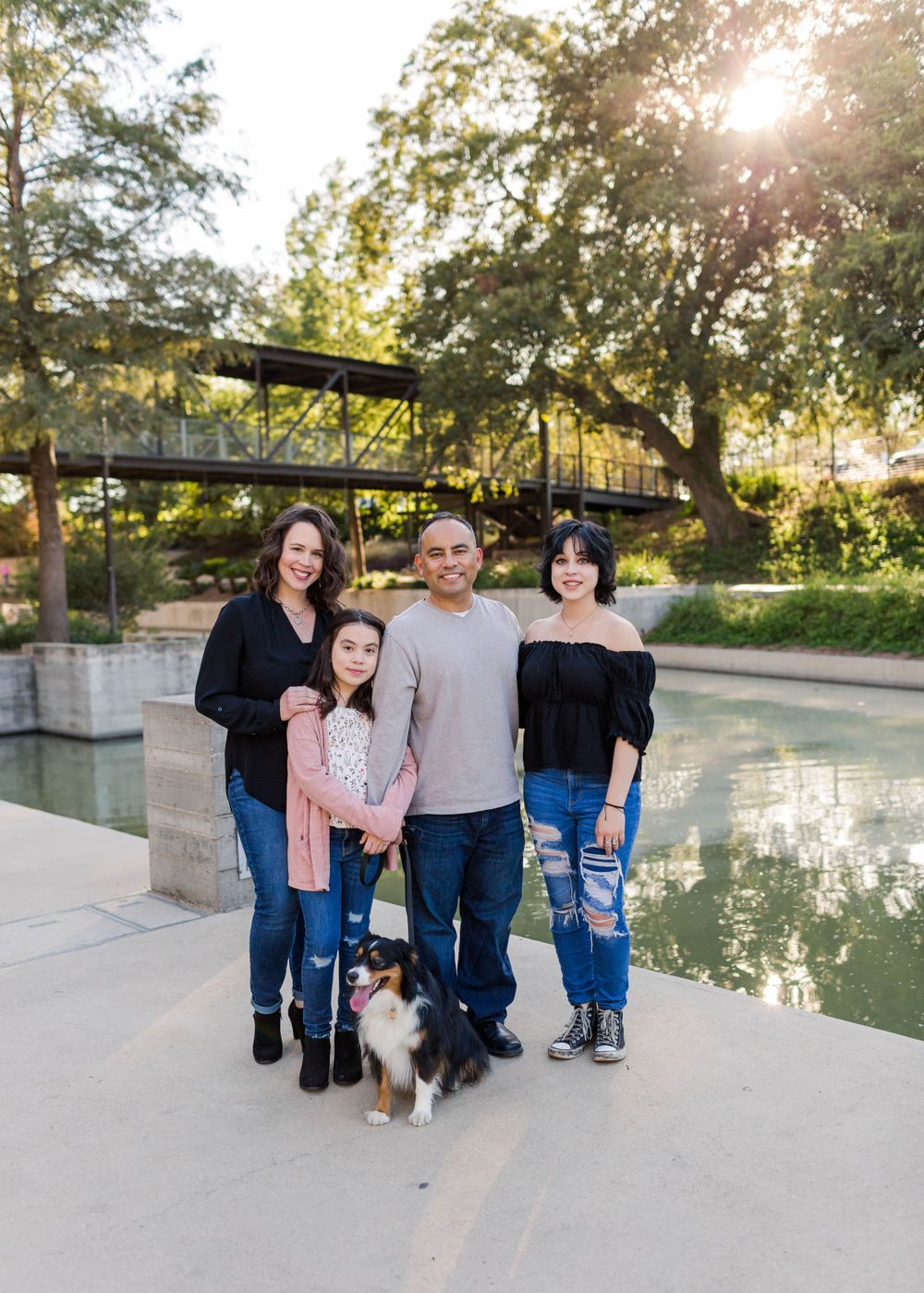 A family and their dog pose together near a reflective pool with a bridge in the background.