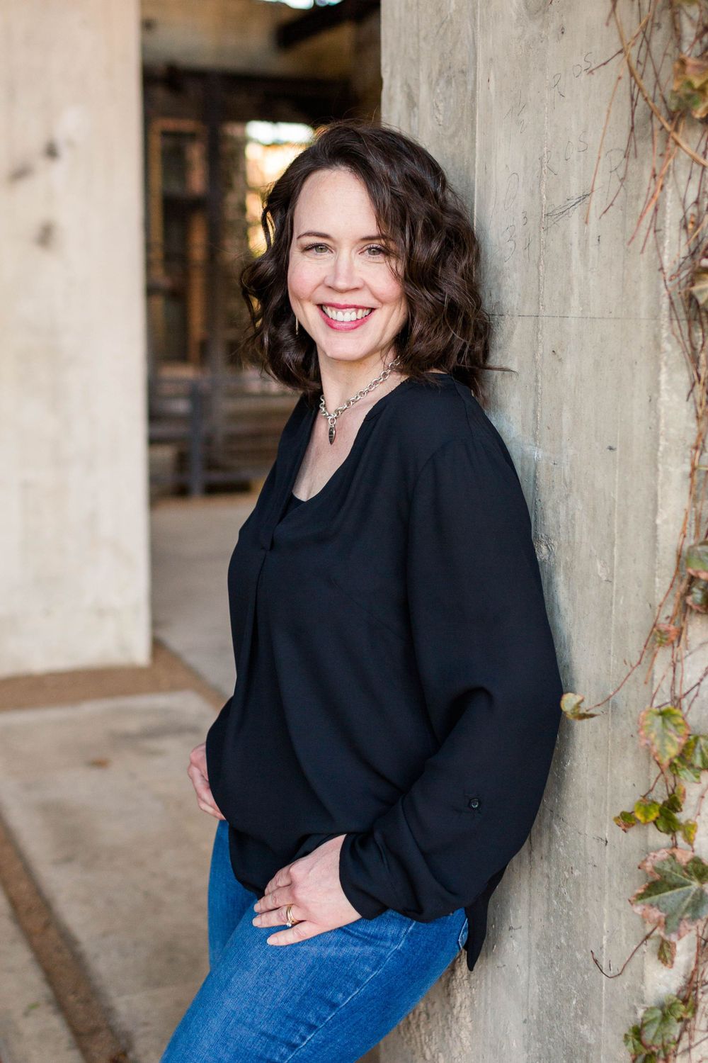 A person in a black sweater leans against a concrete pillar while smiling.
