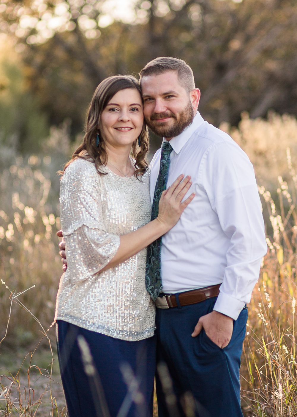 A couple in formal attire poses together in a golden field during sunset.