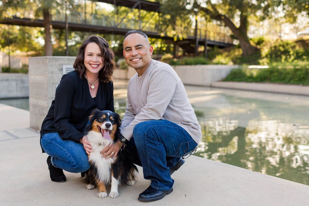 A couple kneels with their small dog by a peaceful waterfront setting.