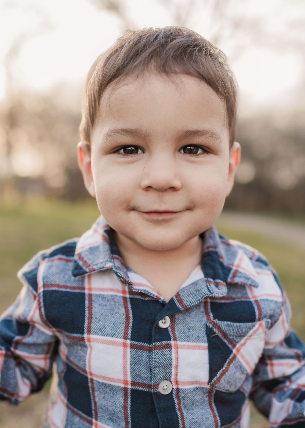 Young child wearing a plaid button-down shirt smiling at the camera outdoors.