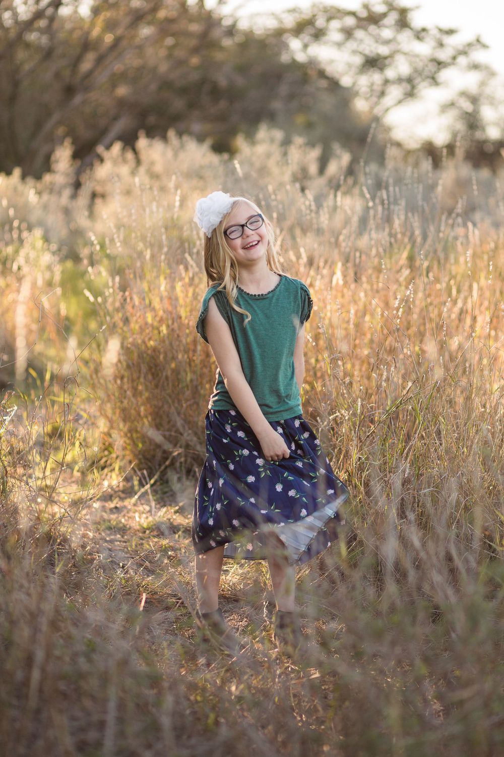 A person in a green shirt and patterned skirt twirls playfully in a sun-lit field of tall grass.