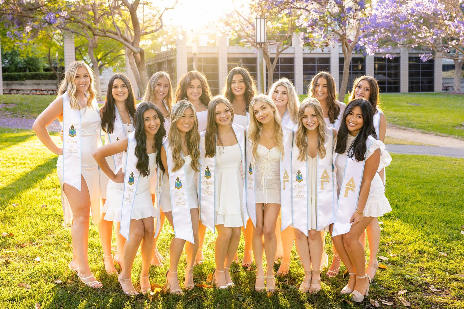 A group of women wearing white dresses pose together on a grassy lawn at sunset.