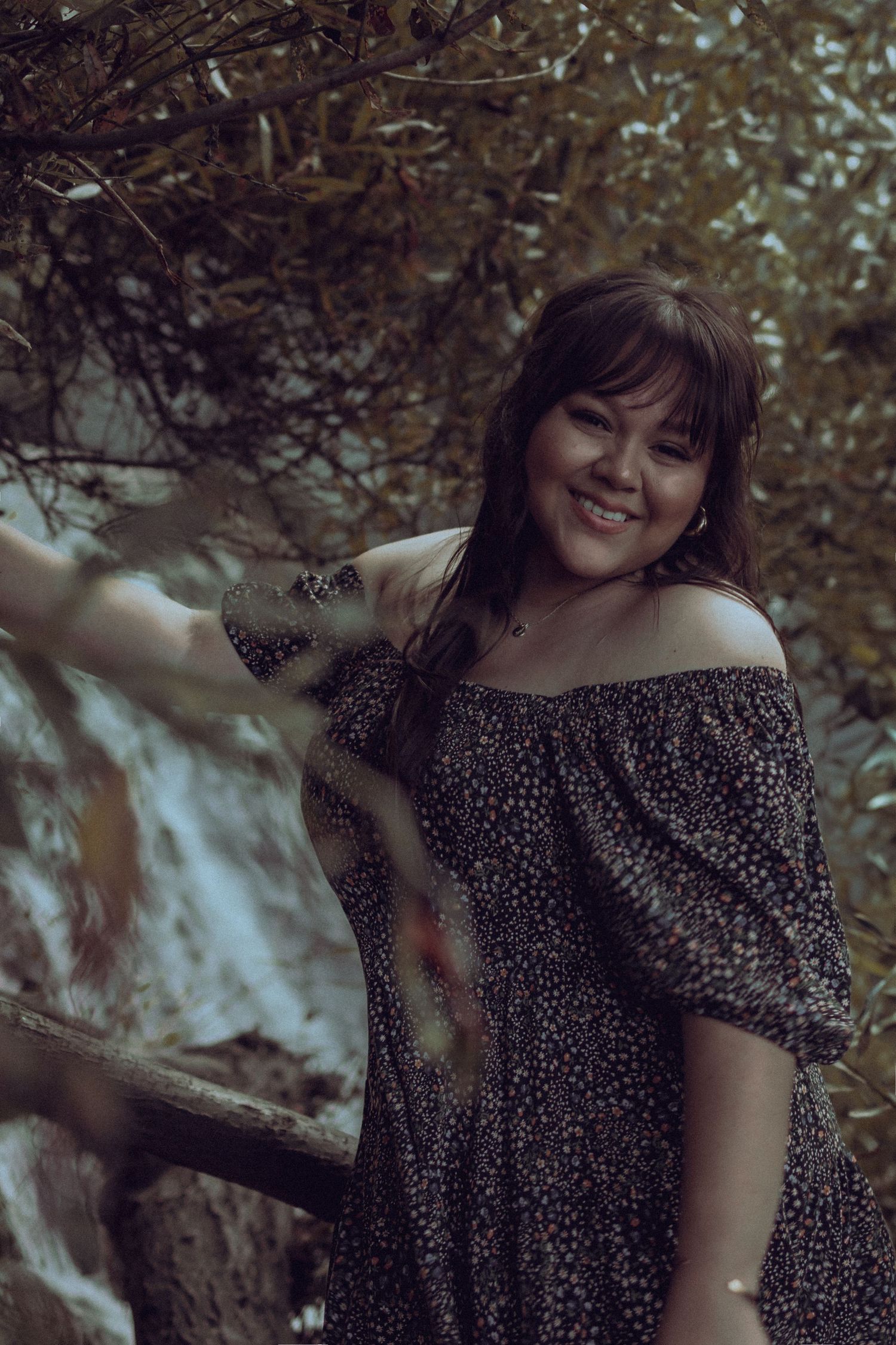Portrait of a smiling woman in an off-the-shoulder floral dress standing in a wooded area.