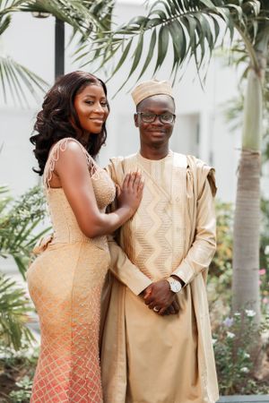 Couple in traditional gold and cream wedding attire posing outdoors near palm trees.