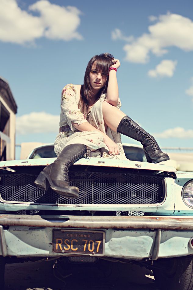 A woman in a lace dress and combat boots poses atop a vintage car with a blue sky background.