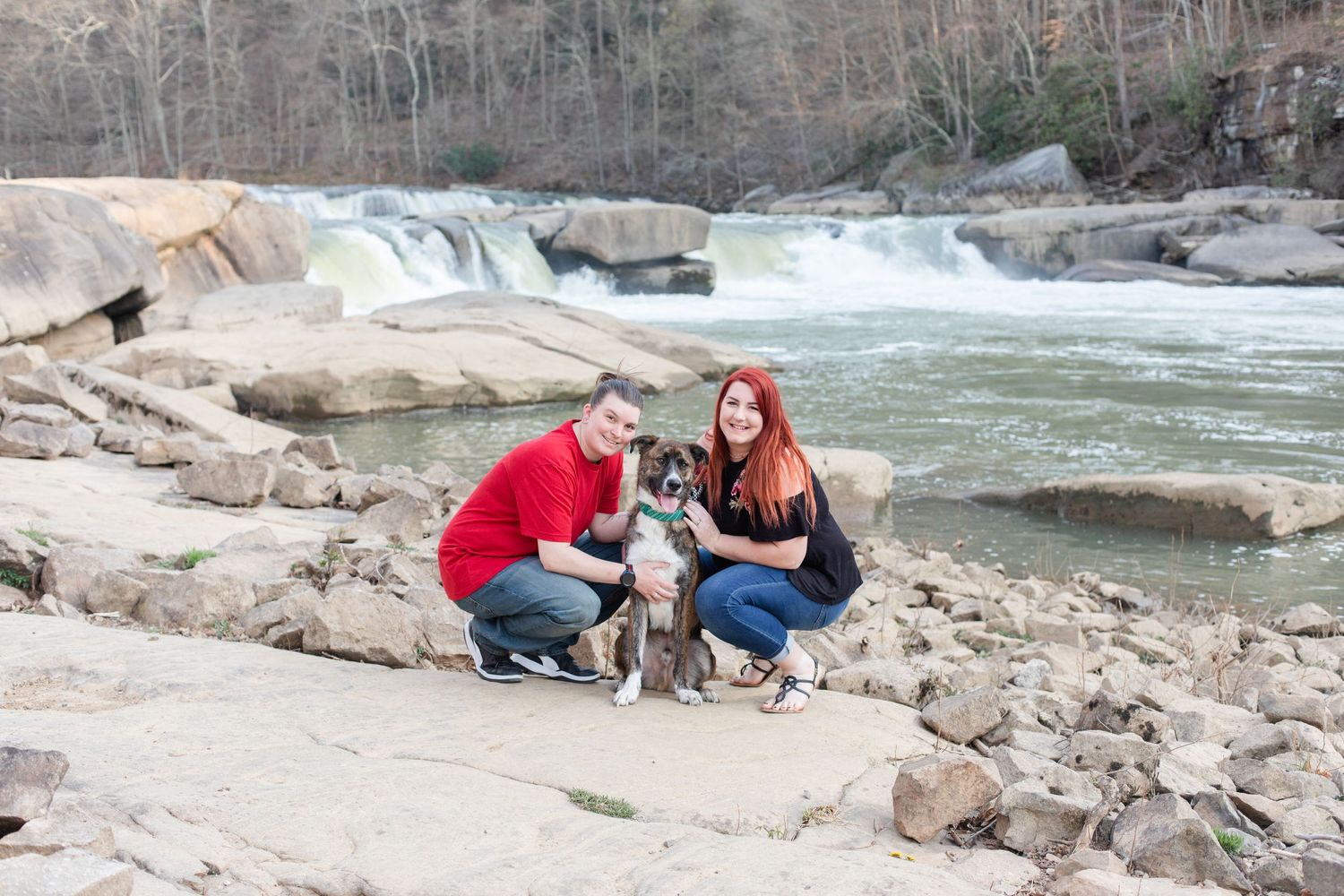 A couple poses with their dog near rushing waters and rocky terrain.
