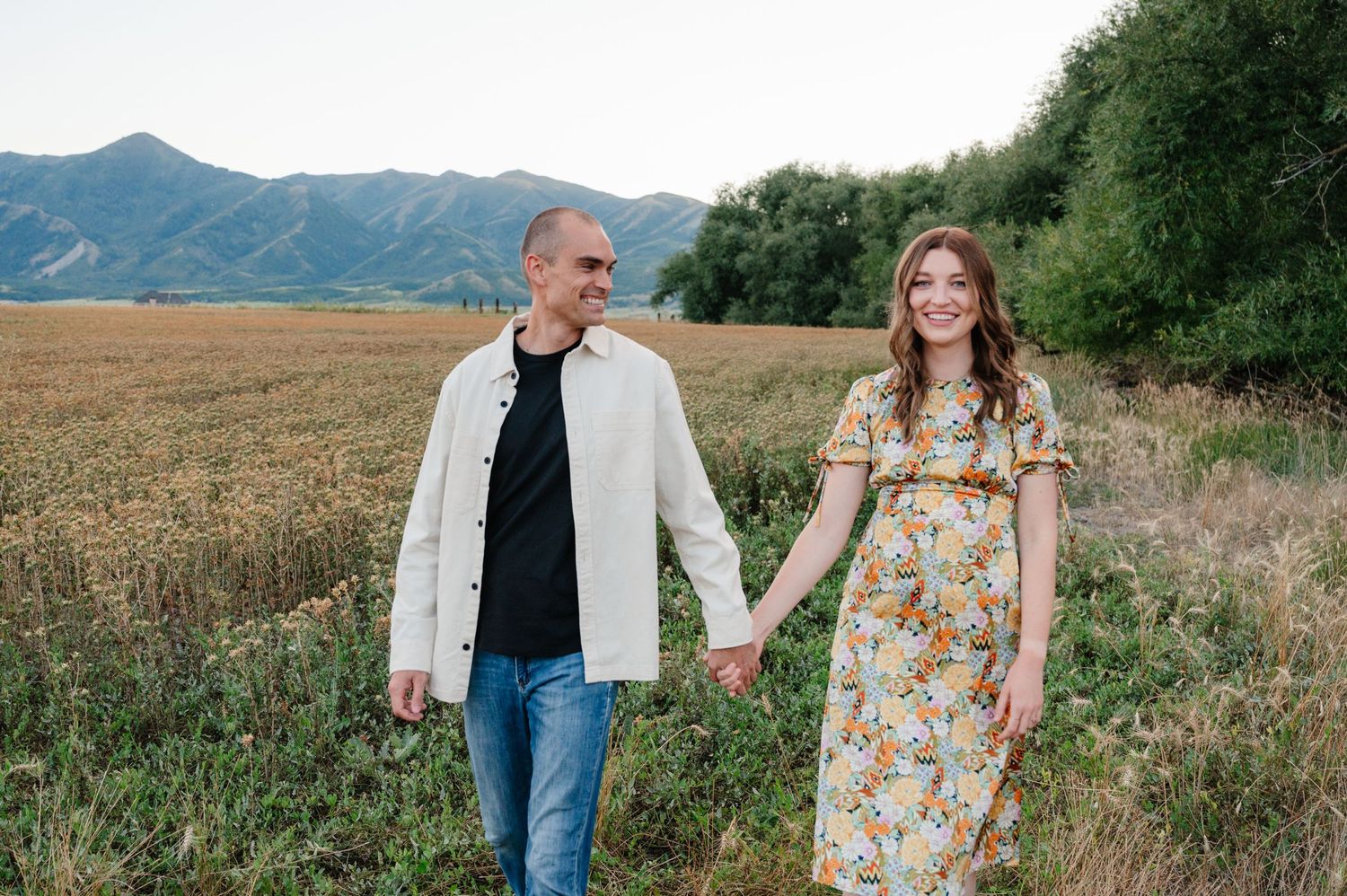 Happy couple holding hands while walking through a summer field with mountain views.