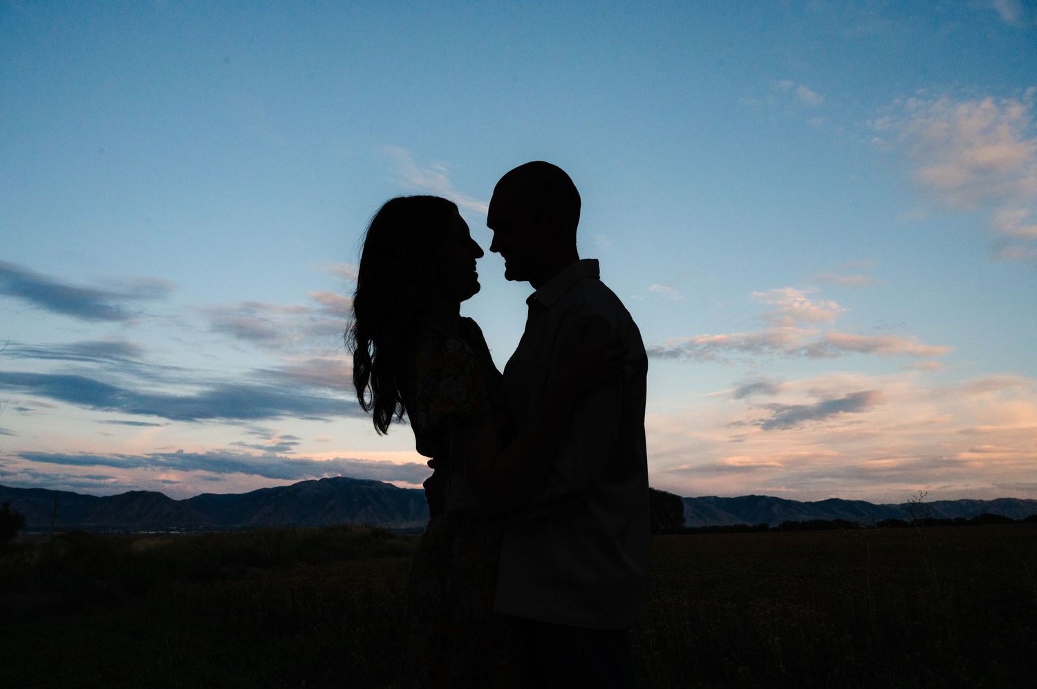 Silhouetted couple in an intimate pose against a dramatic sunset sky with mountains.
