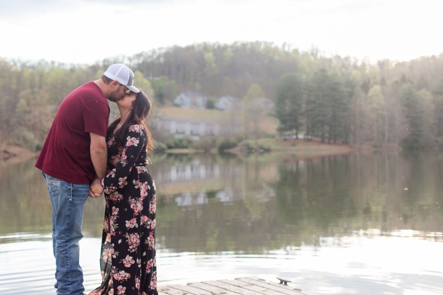 Couple stands close together on wooden pier with tranquil lake and wooded shoreline behind them.