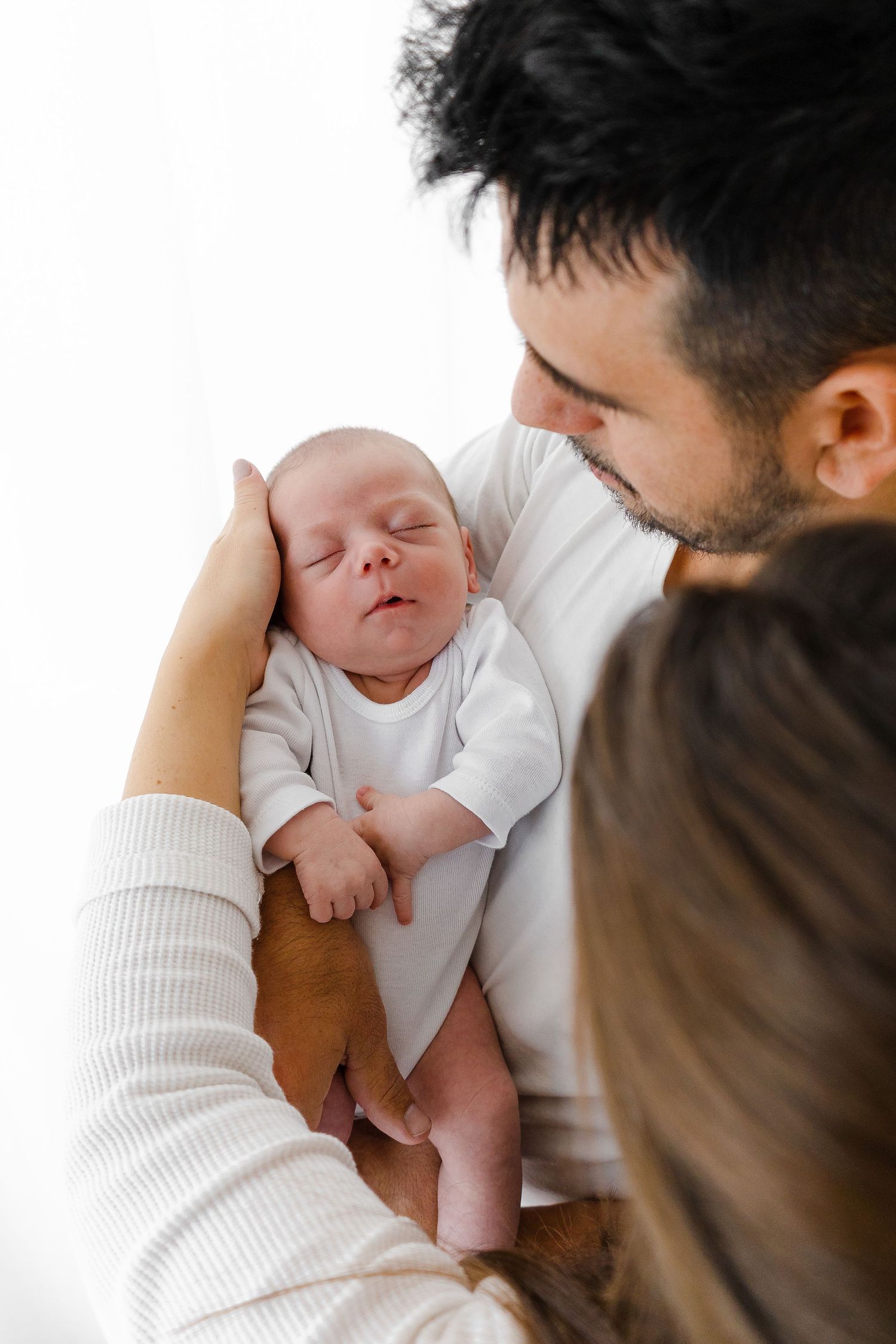 Intimate newborn portrait with parents holding sleeping baby in white onesie.