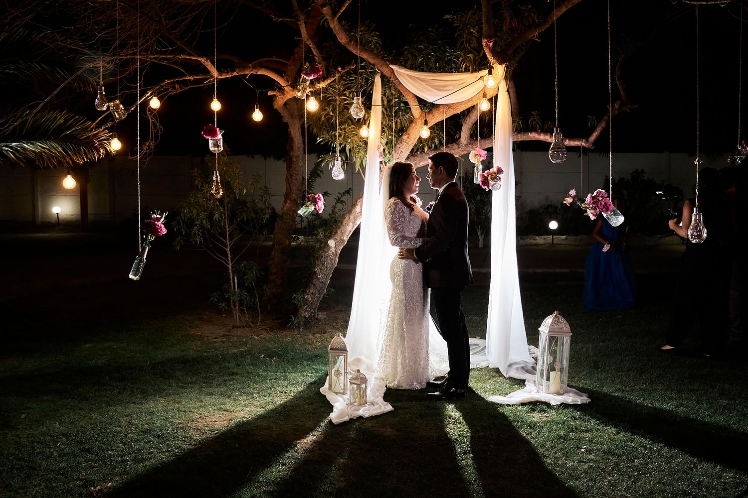 Nighttime wedding ceremony under twinkling lights and draped fabric in an outdoor garden setting.