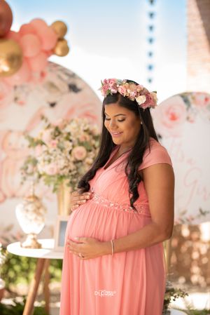 Pregnant woman in pink floral crown and gown cradles baby bump at decorated baby shower event.