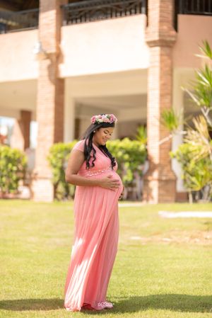 Pregnant woman in pink maxi dress and floral crown cradles baby bump on lush green lawn.