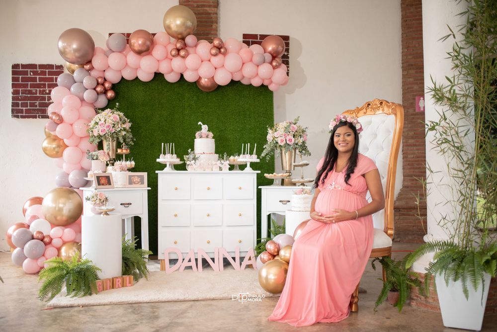Pregnant woman in pink gown sits at elegant pink and gold balloon-decorated baby shower setup.