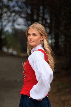 Nordic traditional dress featuring red vest and white blouse against dark woodland background.