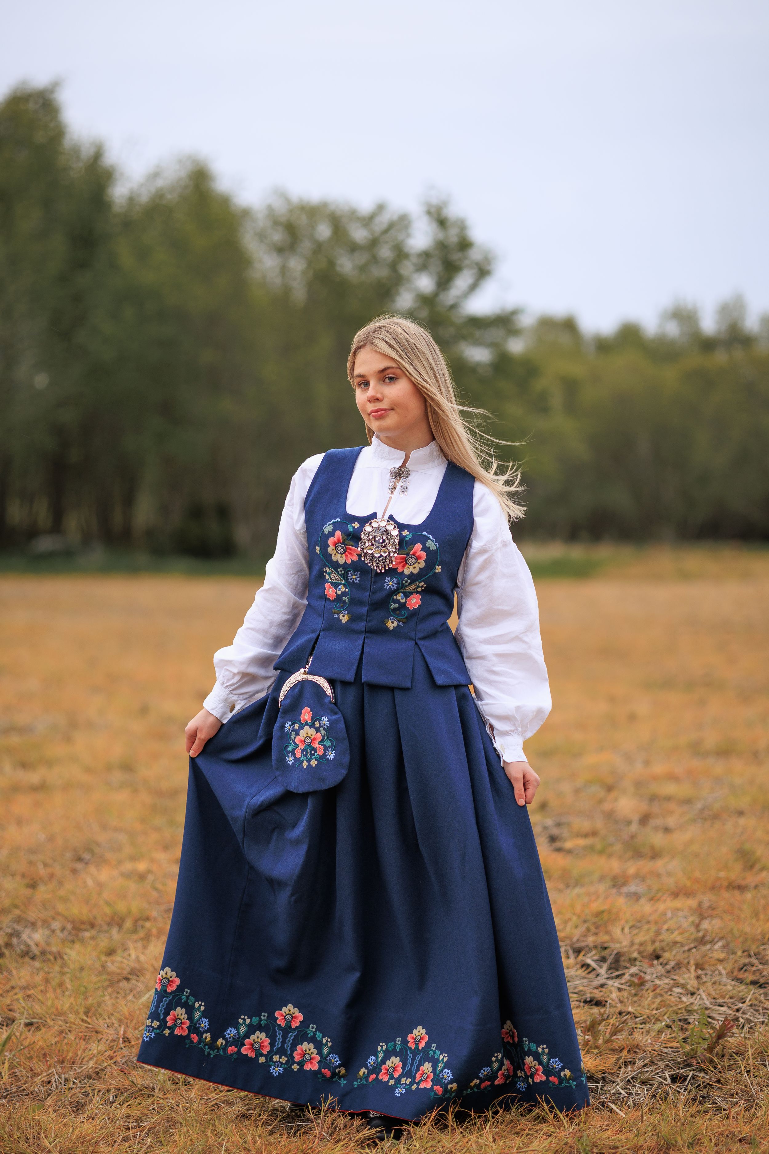 Full-length view of a navy blue Norwegian bunad with floral embroidery in an autumn field.