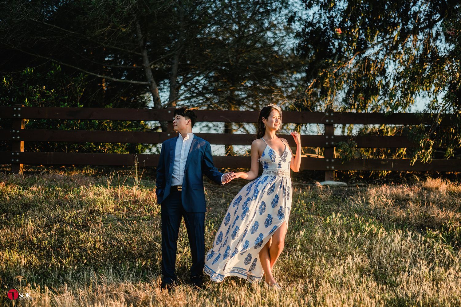 A couple in formal attire poses in a field at sunset near a wooden fence.