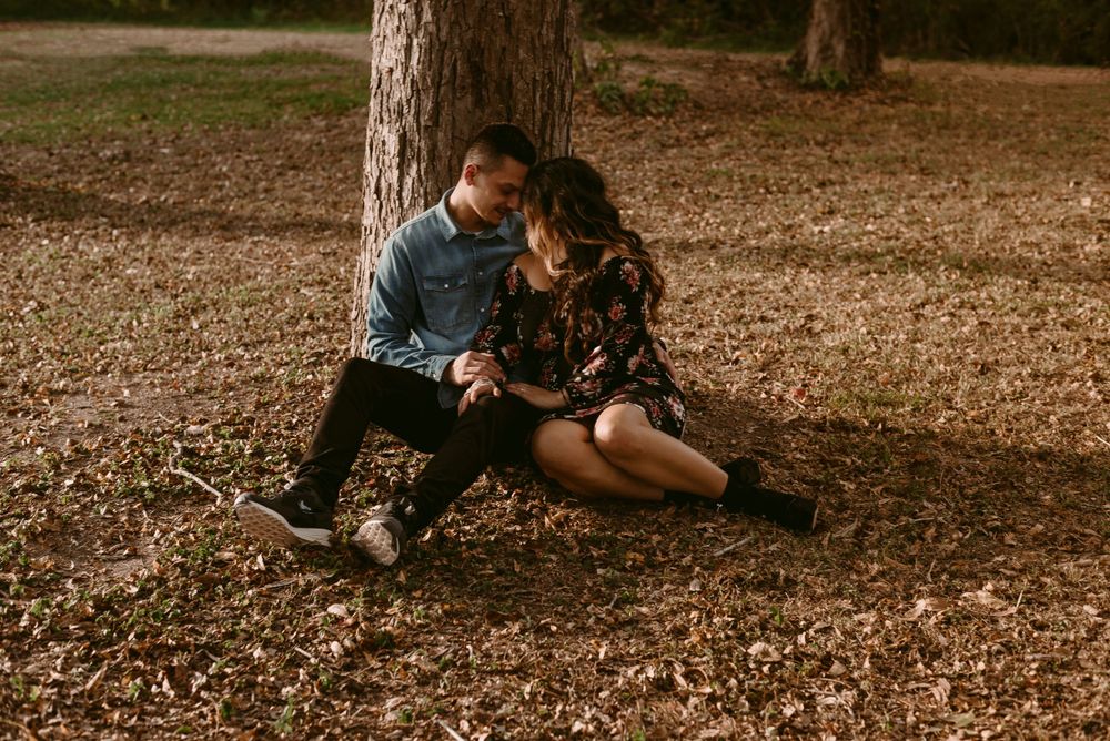 Couple sits together intimately against a tree trunk surrounded by fallen autumn leaves.