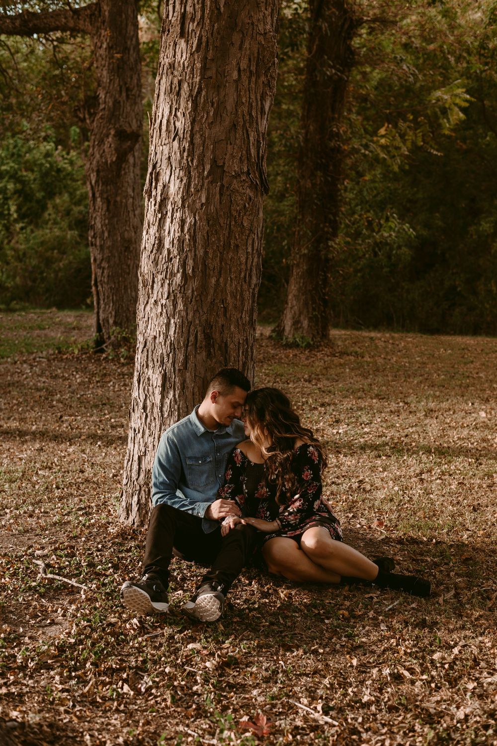 Intimate couple sitting by large tree trunk in romantic woodland setting during golden hour.