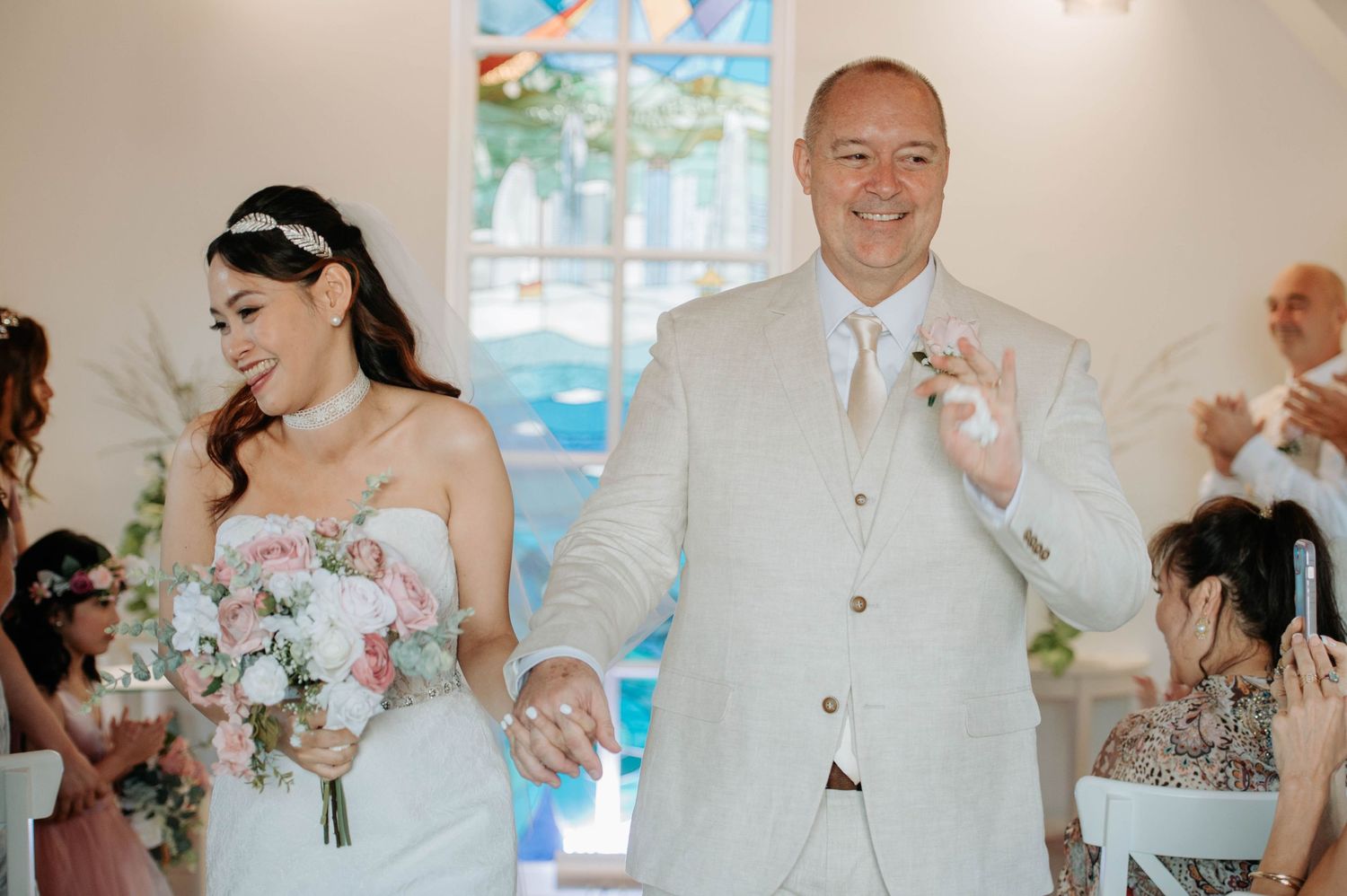 A couple exits their wedding ceremony in a white chapel with a stained glass window.