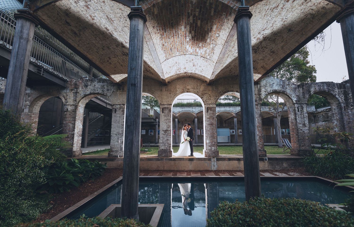 Wedding couple kiss under ornate stone arches reflected in a tranquil pool at historic venue.
