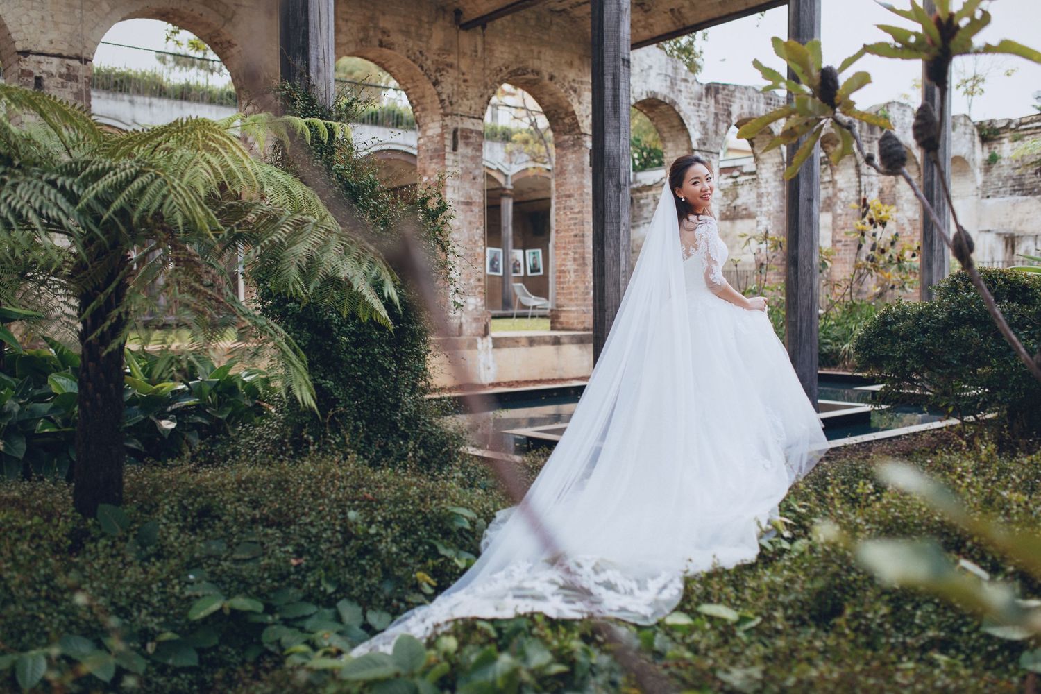 Bride in flowing white gown with long train stands among tropical ferns at ruins venue.