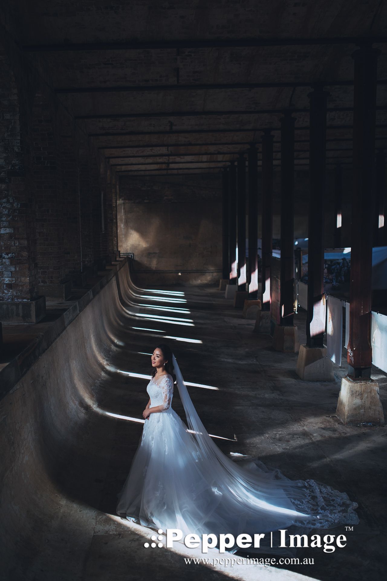 Bride in white gown with veil stands in dramatic light inside historic industrial building.