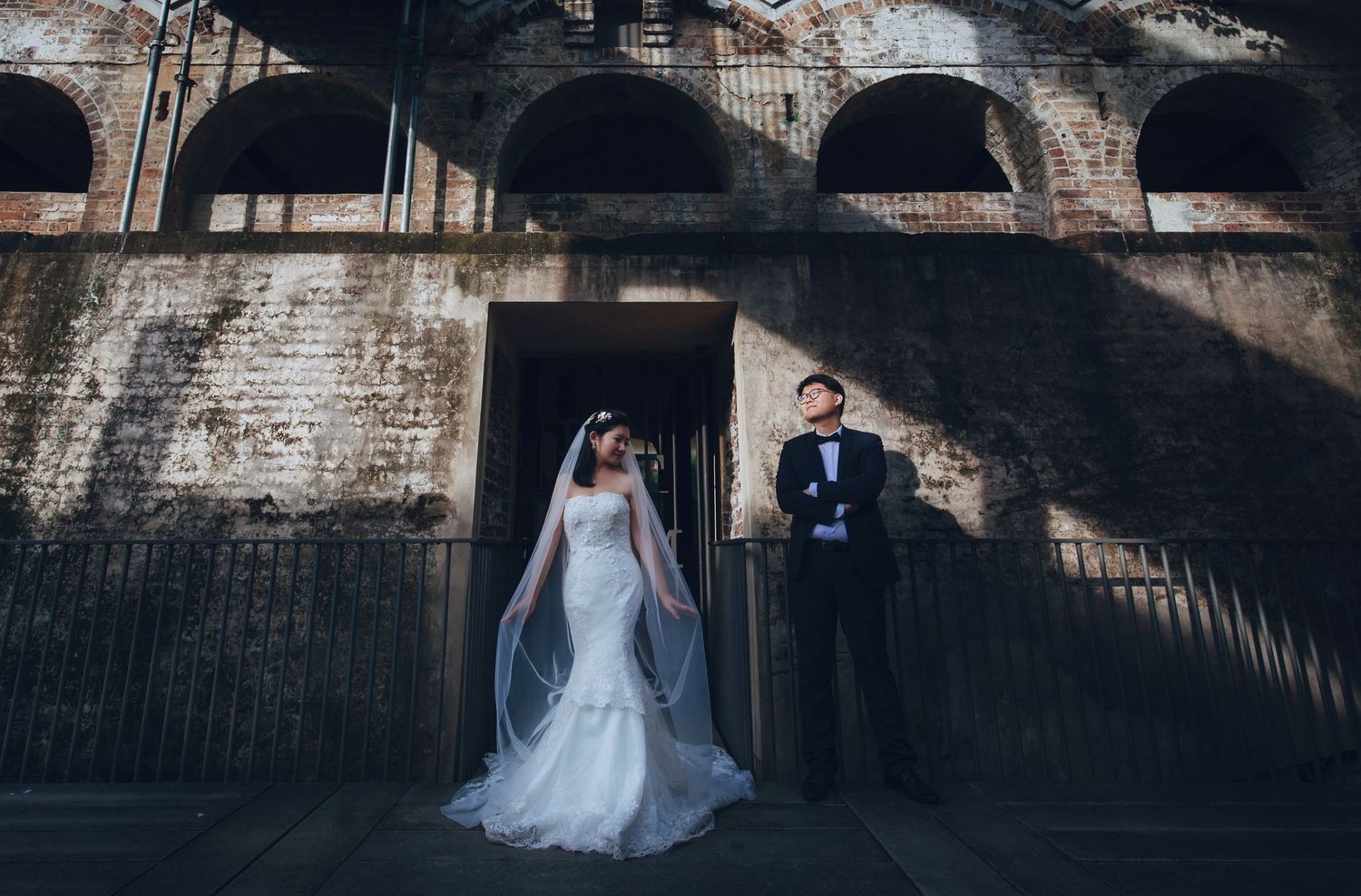 Bride and groom pose dramatically outside historic brick arched building in wedding attire.