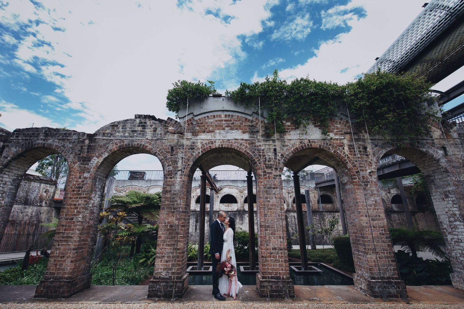 Bride and groom embrace beneath weathered brick arches with lush greenery under blue sky.