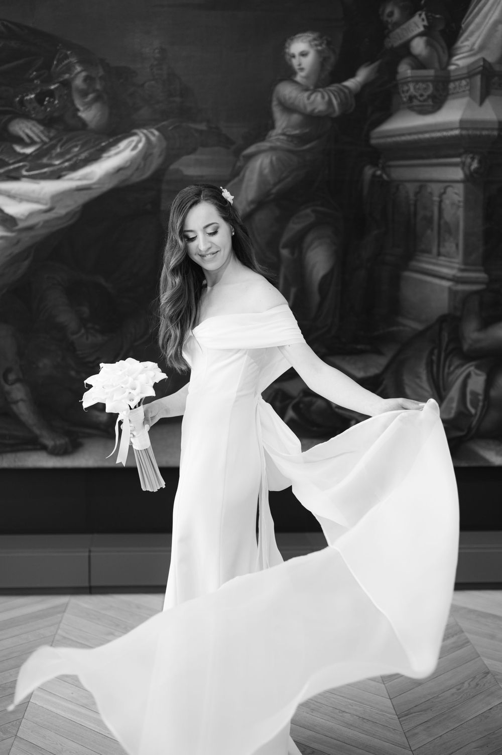A bride in a flowing white long-sleeved gown twirls gracefully while holding a bouquet against an ornate dark background.